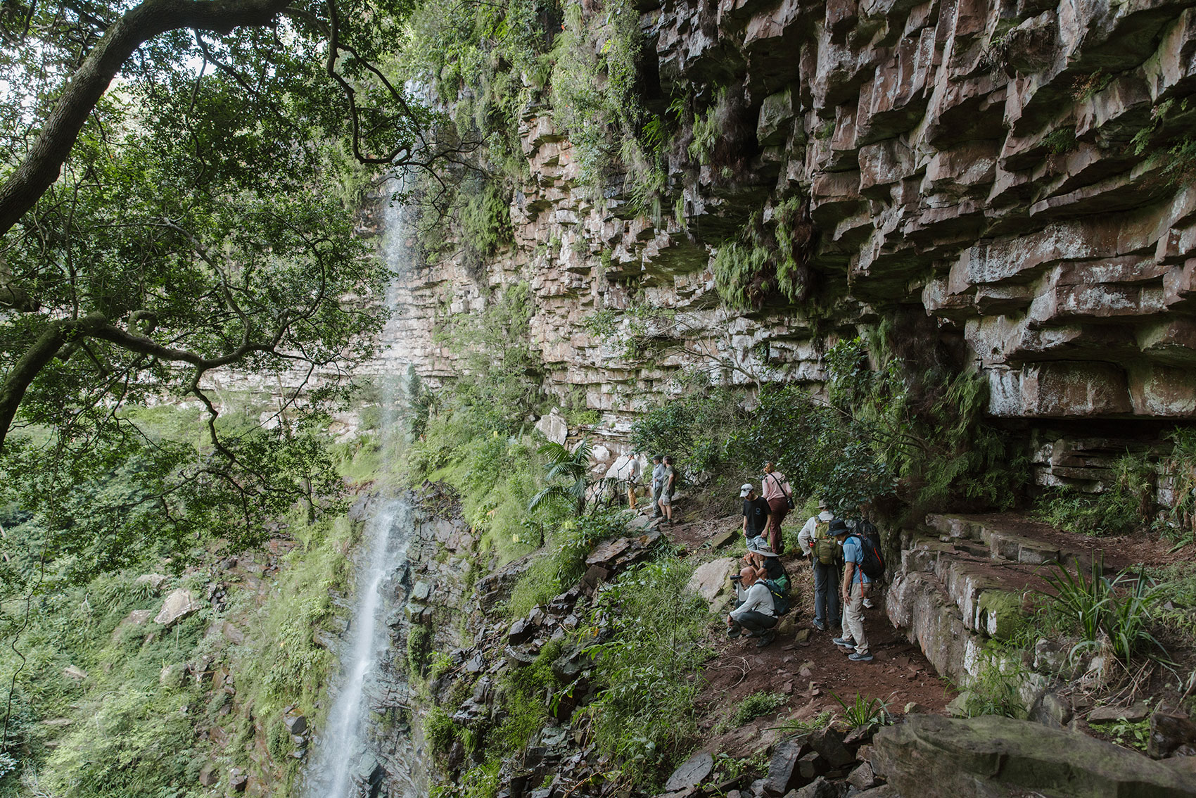 Guests walking beneath a sandstone cliff face beside a waterfall